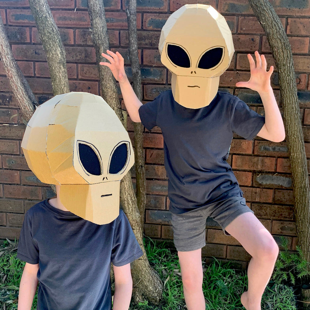 Two children wearing alien head masks in front of a brick wall.