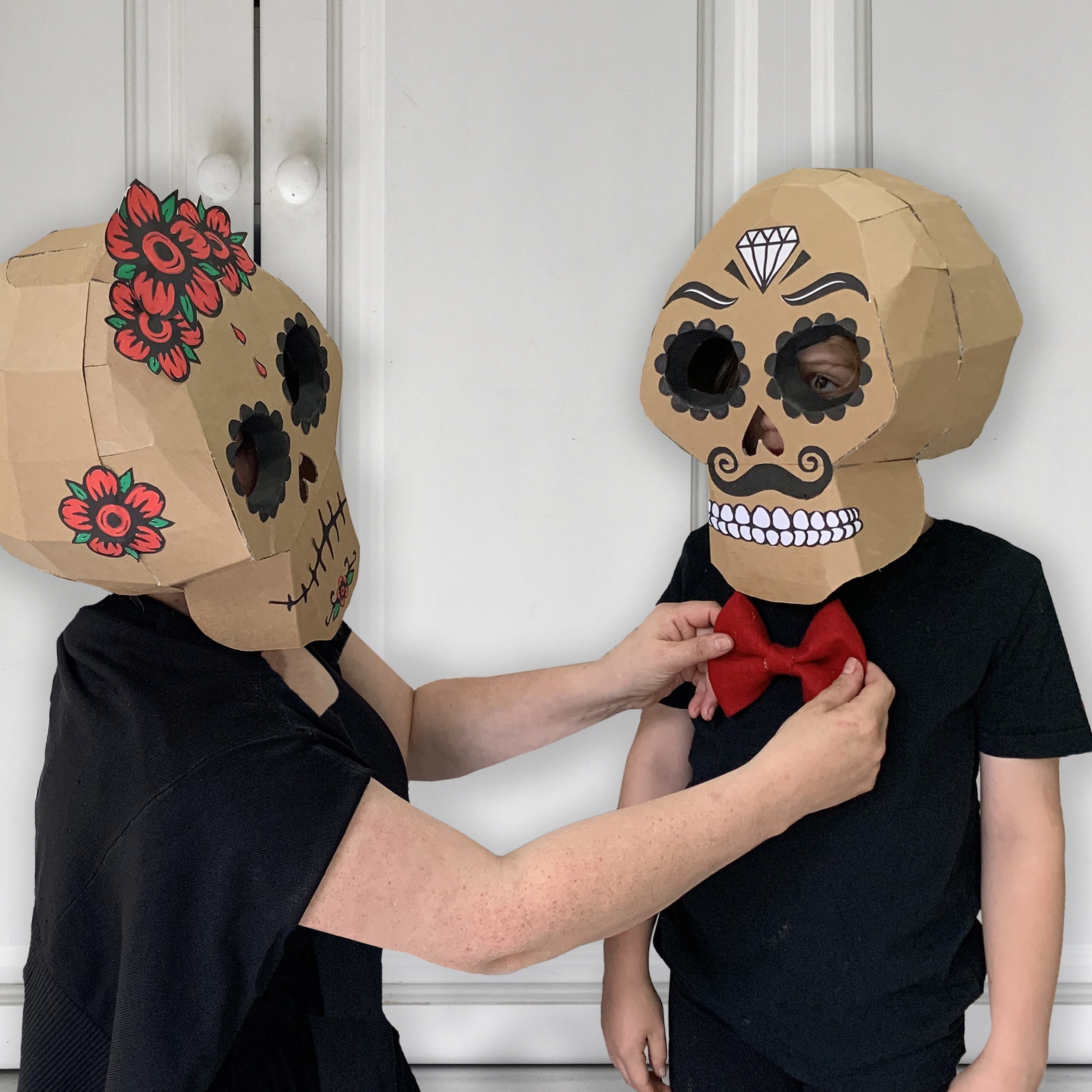 Two people wearing cardboard sugar skull mask costumes with decorative elements against a white wall.
