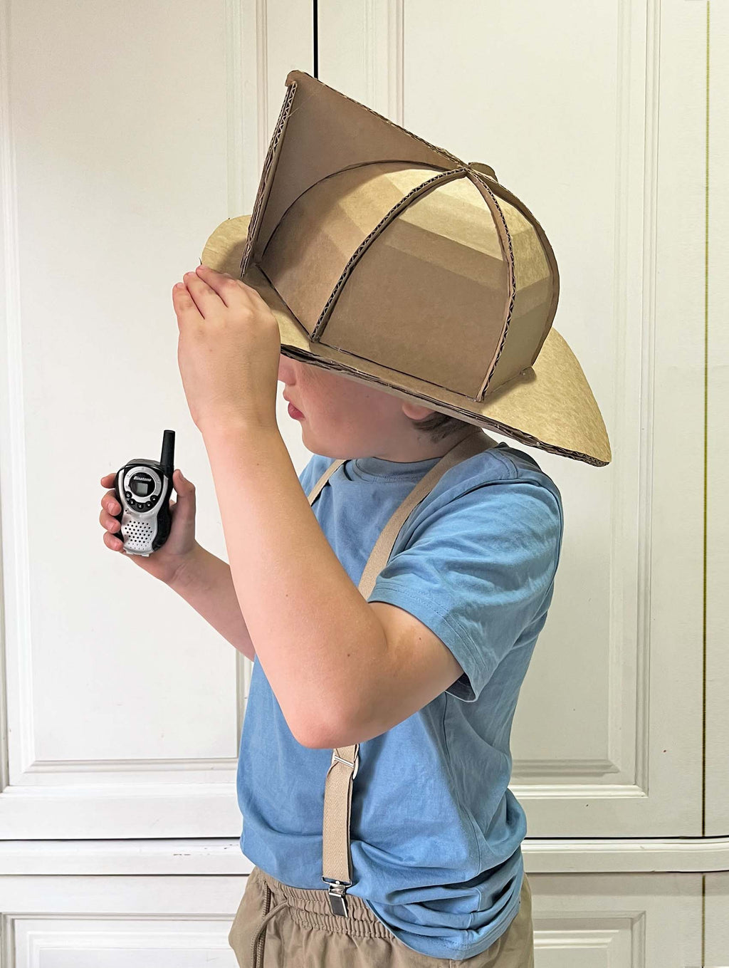 Child wearing a cardboard fireman helmet hat and holding a walkie-talkie indoors.