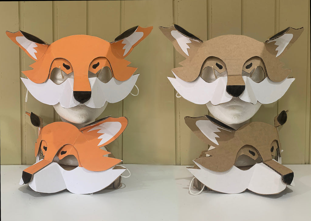 Two cardboard animal masks, one with an orange fox face  and the other with natural cardboard tones. Both using paper overlays over cardboard to create a clever fox mask.
